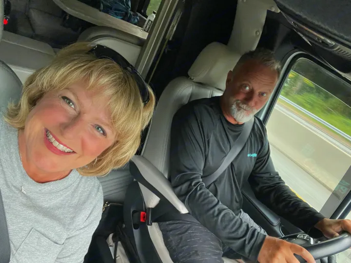 Happy couple in their RV rental, smiling on a road trip. The woman takes a selfie from the passenger seat, man drives.. Thor Tiburon 2020