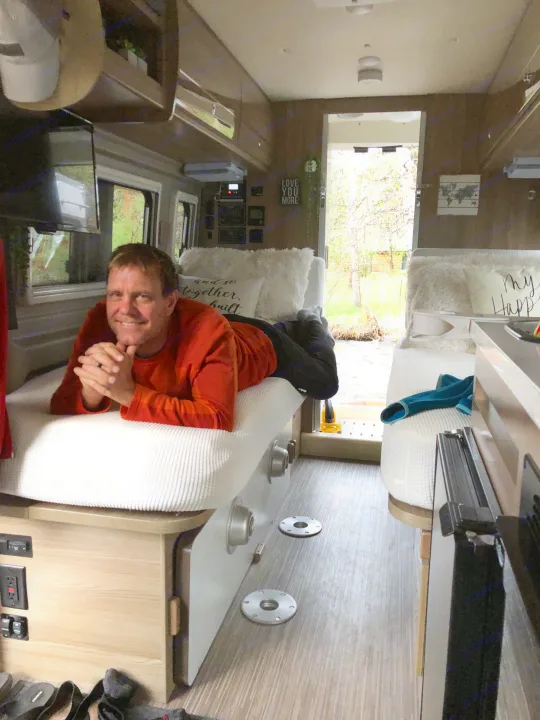Man smiles from a comfortable bed in an RV rental, overlooking a scenic outdoor view. Ideal for travel and relaxation.. Winnebago Travato 2019