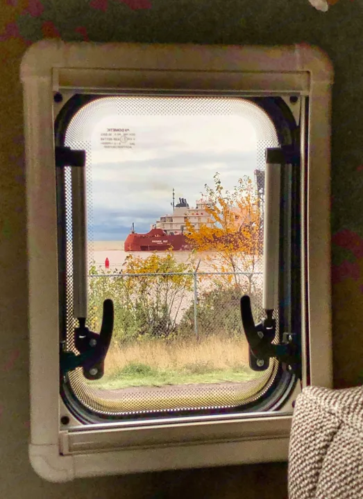 Scenic RV rental view: The red 'Presque Isle' ship on water, framed by an RV window with autumn foliage.. Winnebago Travato 2019