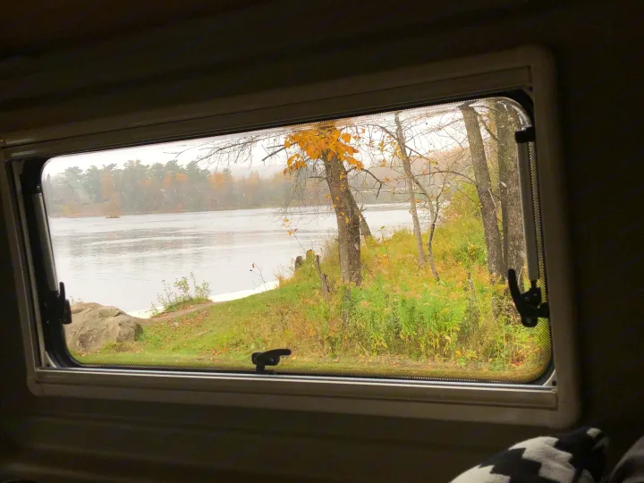 Scenic RV rental window view of a quiet river, autumn foliage, and falling snow on a cloudy day.. Winnebago Travato 2019