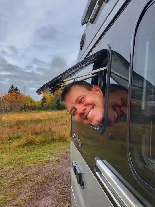 Smiling man and his reflection peek from an RV rental window in a vibrant fall landscape.. Winnebago Travato 2019