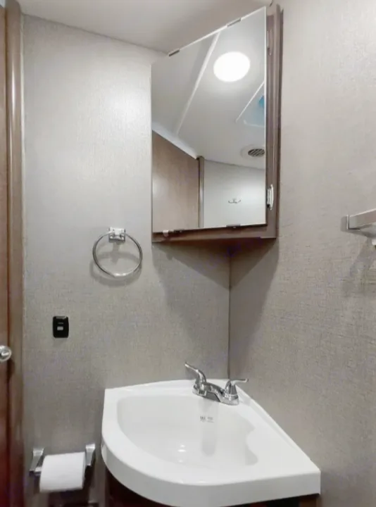 Compact RV rental bathroom featuring a corner sink, medicine cabinet, towel ring, and a mirrored reflection of the ceiling light and vent.. Winnebago Outlook 2019