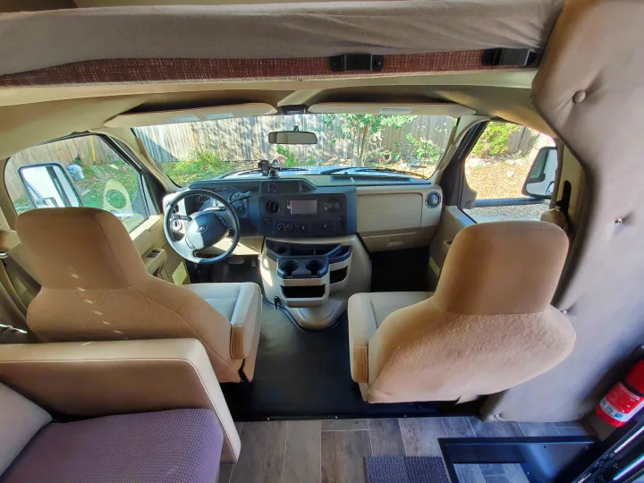 RV rental interior view: Spacious front cockpit with tan driver & passenger seats, dashboard, and overhead bunk.. Winnebago Outlook 2019