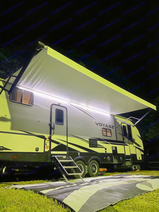 Nighttime RV rental scene. Illuminated modern RV with extended awning, steps, and mat on grass. Perfect for camping.. Winnebago Voyage 2021