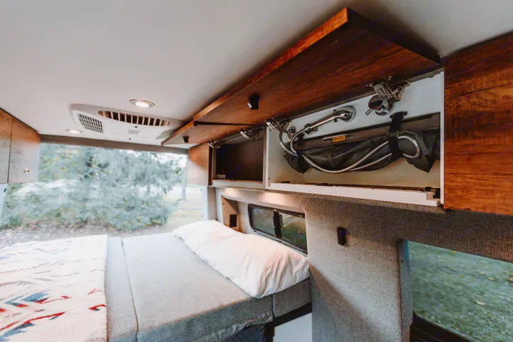 View of an RV rental interior: comfortable bed, overhead cabinets with a stowed portable shower, and window view.. Story Teller Classic Mode 4X4 2022