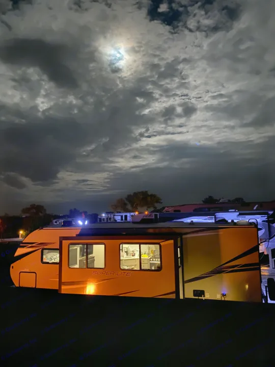 Night view of an RV rental: a bright travel trailer illuminated under a moonlit, cloudy sky at a campsite.. Starcraft Super Lite 2022