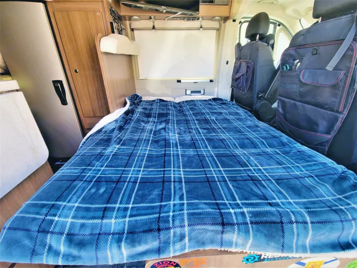 Cozy RV rental interior with a blue plaid bed, fridge, wooden cabinets, and front seat organizers, ready for travel.. Winnebago Travato 2018
