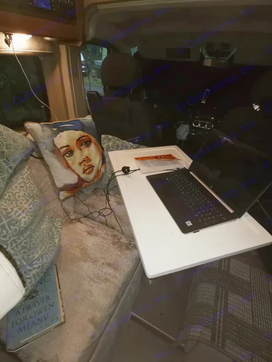 Cozy RV rental interior setup: laptop on a portable desk, 'A Prayer for Owen Meany' book, and comfy pillows.. Winnebago Travato 2018