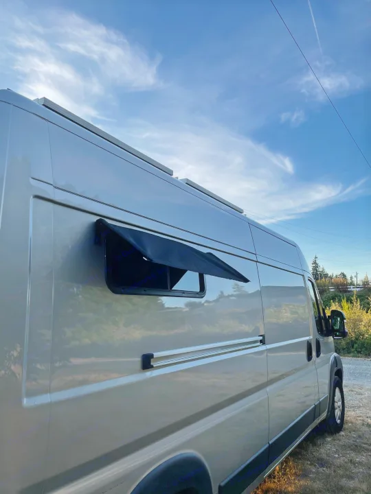 Side view of a modern RV rental camper van, light grey with solar panels and a window awning, parked outdoors under a blue sky.. Dodge Other 2018