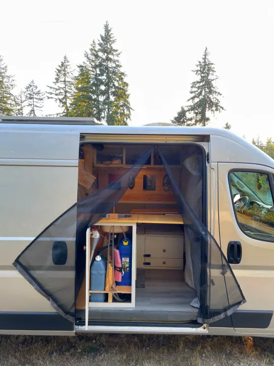 Spacious camper van for rv rental, showing open side door with bug screen, interior sink, and bed. Trees in background.. Dodge Other 2018
