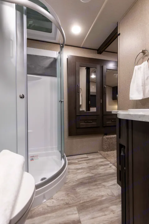 Modern RV rental bathroom with a curved glass shower, dark wood vanity, and mirrored storage cabinet. Toilet visible foreground.. Grand Design Reflection 2021