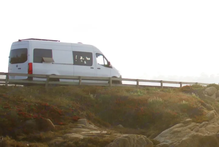 A white camper van with an open side vent is parked on a scenic road by a fence, overlooking rocky coastal terrain. Great RV rental option.. Mercedes-Benz Sprintervan 2019