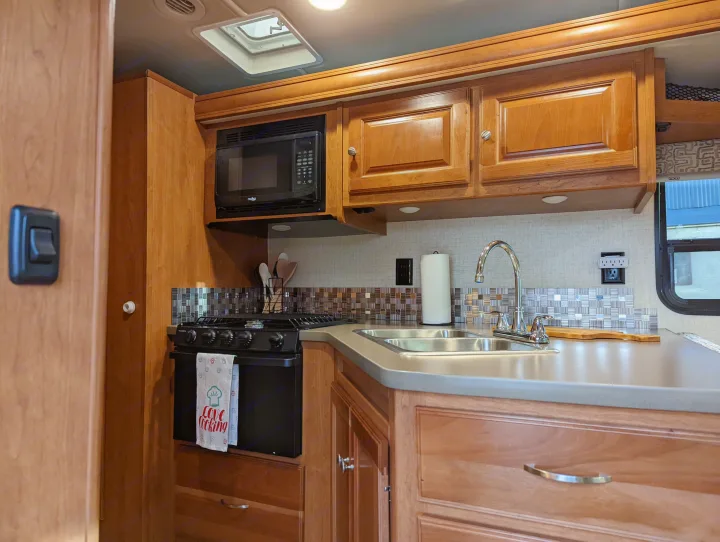 Compact RV rental kitchen with microwave, stove, double sink, and wooden cabinets.. Winnebago Vista 2018