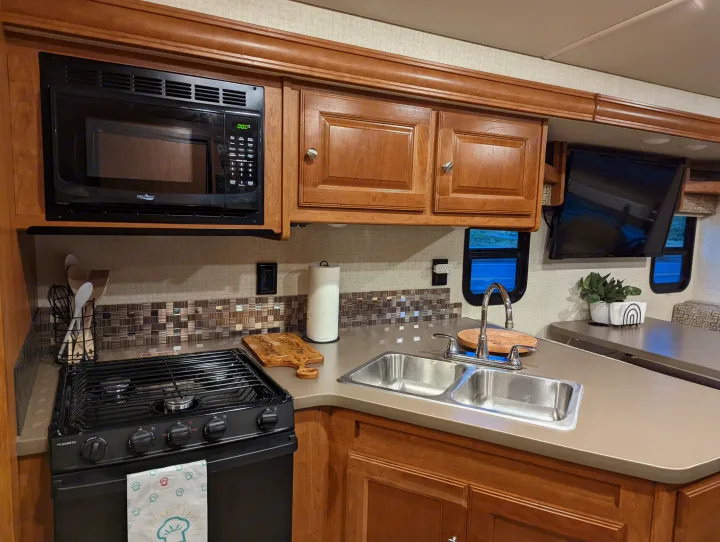 A well-equipped rv rental kitchen featuring a microwave, stove, double sink, wood cabinets, and dining area with TV.. Winnebago Vista 2018