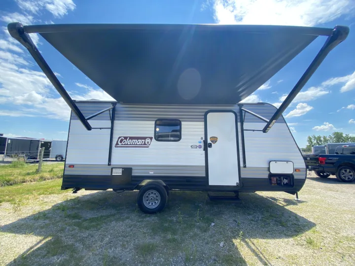 A Coleman travel trailer, ready for RV rental, with its awning extended under a sunny sky. Parked on a gravel lot.. Dutchmen Coleman Lantern LT 2023