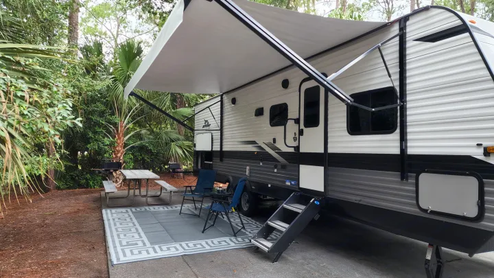 RV rental parked at a shady campsite. An extended awning covers an outdoor rug, chairs, and a picnic table amidst lush greenery.. Jayco Jay Flight SLX 2022