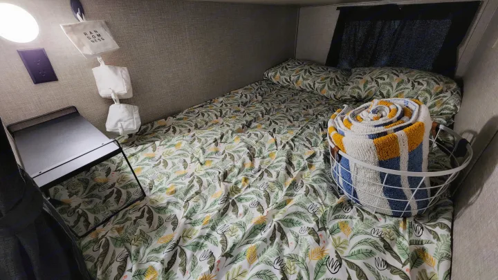 Cozy RV rental bunk with leafy bedding, a small shelf, hanging storage bags, and a basket of striped towels.. Jayco Jay Flight SLX 2022