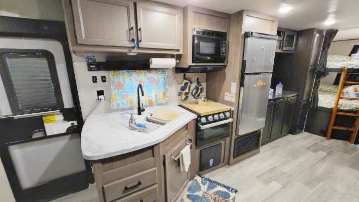 Bright RV rental kitchen on the left with sink, microwave, fridge; bunk beds on the right. Modern wood-look interior.. Jayco Jay Flight SLX 2022