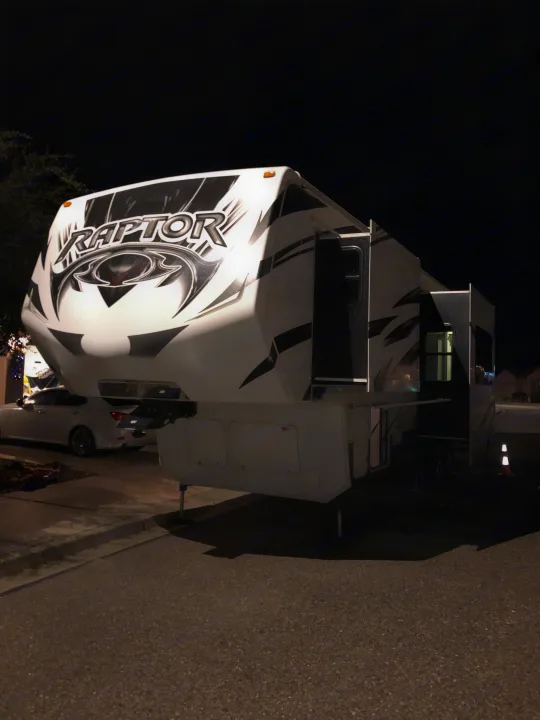A large white and black Raptor fifth-wheel RV is parked at night with slide-outs extended. Perfect for an RV rental.. Keystone Raptor 2014