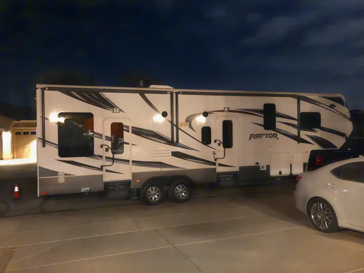 Night view: Large white Raptor RV rental, a fifth wheel, with black graphics, parked on driveway. Ready for travel.. Keystone Raptor 2014