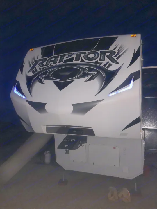 Night shot of a white Raptor fifth-wheel RV, perfect for rv rental adventures, featuring distinctive blue LED lights on its front cap.. Keystone Raptor 2014