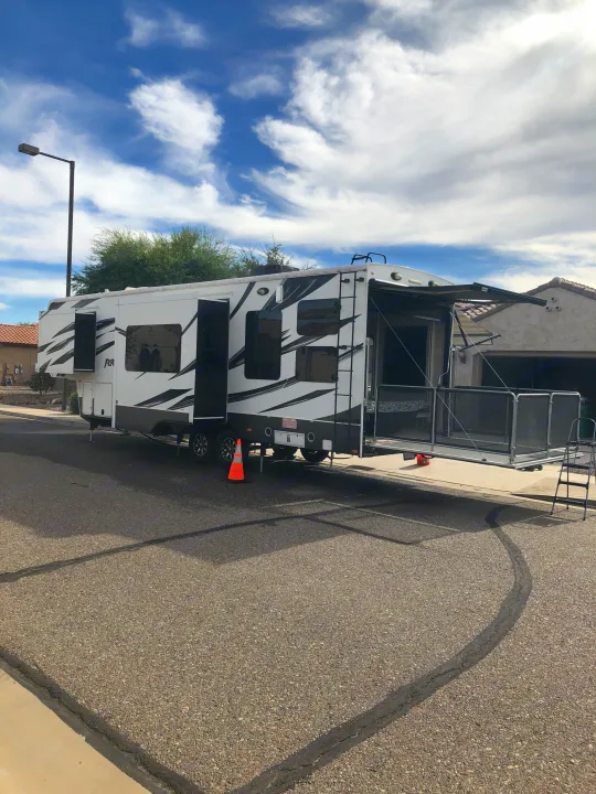 A large white RV rental with black graphics is parked on an asphalt street. Its rear ramp is down, forming a patio with an awning extended.. Keystone Raptor 2014