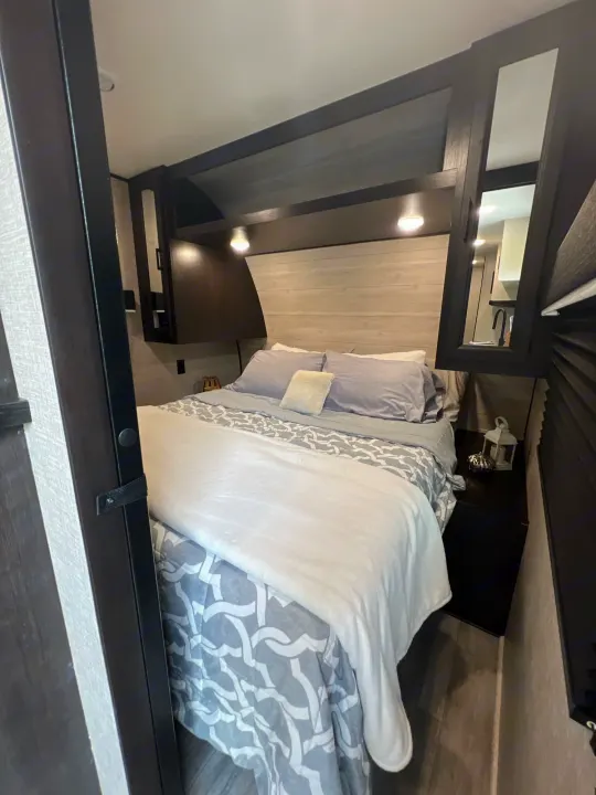 Step inside this modern RV rental bedroom, featuring a comfy bed with grey bedding, built-in storage, and task lighting.. Jayco Jay Flight 267BH 2023