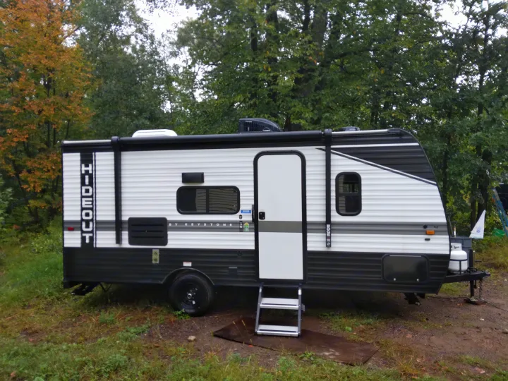A white and black Keystone Hideout travel trailer, an ideal RV rental, is parked outdoors among trees.. Keystone RV Hideout 2022