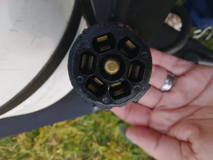 Close-up: A hand holds a 7-pin RV electrical connector, essential for an RV rental trailer. Worn black plug with brass pins.. Keystone RV Hideout 2022