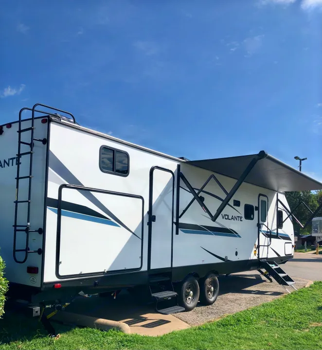 White travel trailer RV rental with an extended awning and ladder, parked on gravel under a clear blue sky.. Crossroads RV Volante 2022