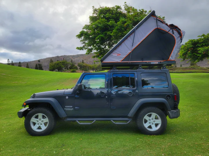 This dark grey Jeep Wrangler, equipped with an open rooftop tent, is ready for an RV rental camping trip on lush green terrain.. Jeep Wrangler 2017