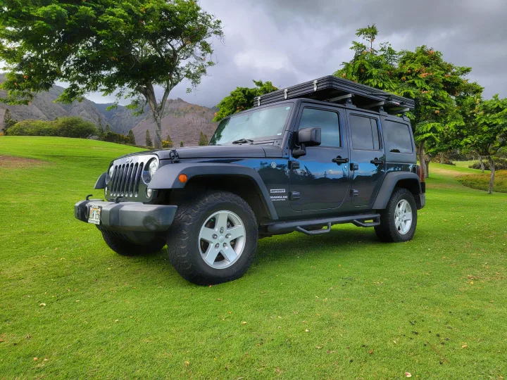 Dark blue Jeep Wrangler with rooftop tent on green grass. Mountains and trees in background. Ideal RV rental.. Jeep Wrangler 2017