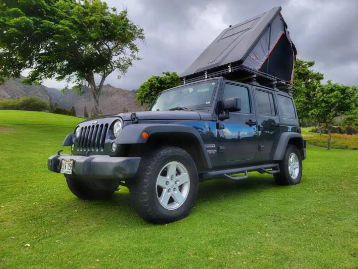 A black Jeep Wrangler with a rooftop tent, ideal for RV rental, sits on green grass with mountains in the background.. Jeep Wrangler 2017
