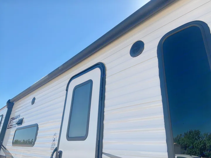Exterior side of a white Coleman rv rental, showing its corrugated design, windows, and door under blue sky.. Dutchmen Coleman Lantern 2021