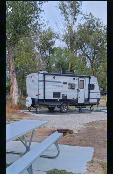 A white Viking RV rental travel trailer is parked on a gravel pad at a campsite, with a picnic table and fire pit nearby.. Forest River Viking 2018
