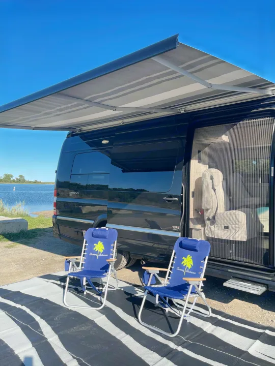 Black Airstream Interstate rv rental with awning. Two Tommy Bahama chairs & mat by a lake. Relaxing outdoor setup.. Airstream Interstate 2018