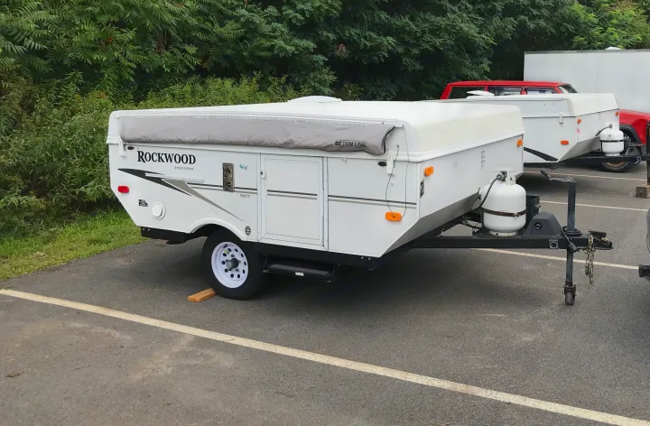 This white Rockwood pop-up camper, equipped with a propane tank and parked on asphalt, is an ideal RV rental for outdoor adventures.. Forest River Rockwood LTD 2010