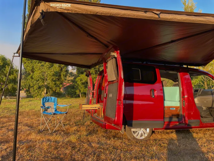 A red camper van RV rental with an open side, revealing a custom interior, parked under an awning with a chair outside.. Nissan Elgrand 2021