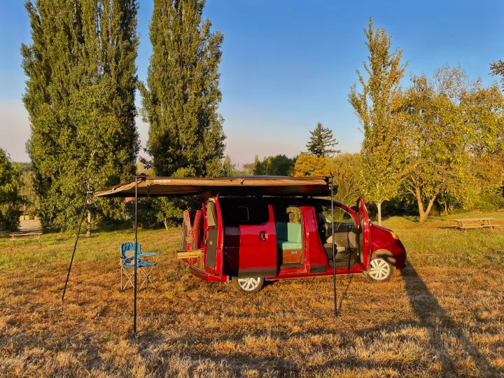 Red camper van RV rental set up with an awning and chair in a sunny field, ready for outdoor adventure.. Nissan Elgrand 2021