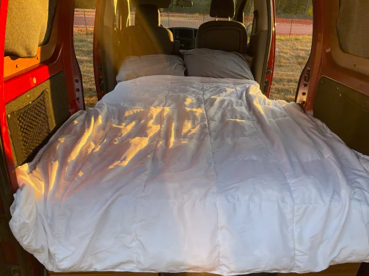 Experience comfort in this cozy rv rental! Van interior with a made bed, white comforter, and sunlit pillows.. Nissan Elgrand 2021