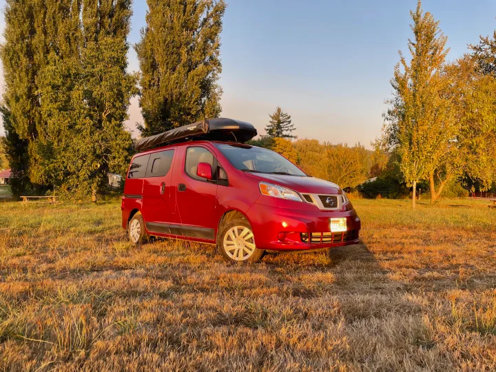 Red Nissan NV200 camper van, possibly an RV rental, parked in a sunny field with trees. Ideal for travel and camping.. Nissan Elgrand 2021
