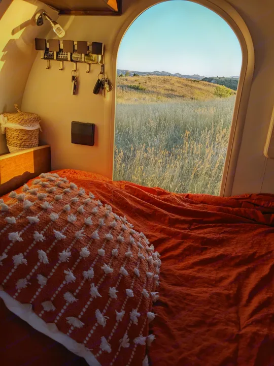 Cozy RV rental bedroom with a sunlit view of a grassy field and distant hills. Keys hang on the wall.. T@G XL 2016