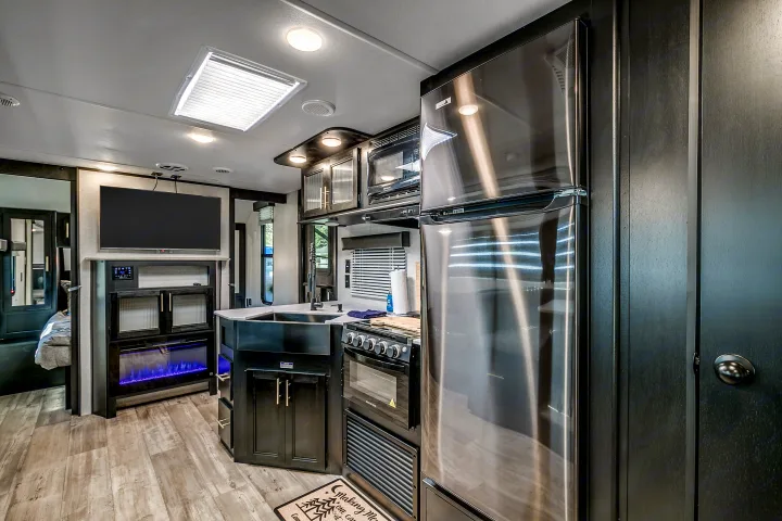 Modern RV rental interior: dark wood kitchen, stainless steel fridge/stove, TV, and blue electric fireplace. Bright and sleek.. Cherokee Grey Wolf 2022