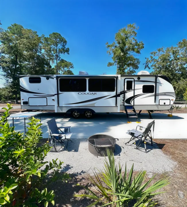 A bright day at an RV rental campsite featuring a Cougar travel trailer, outdoor seating, and a fire pit setup.. Keystone Cougar Half-Ton 2021
