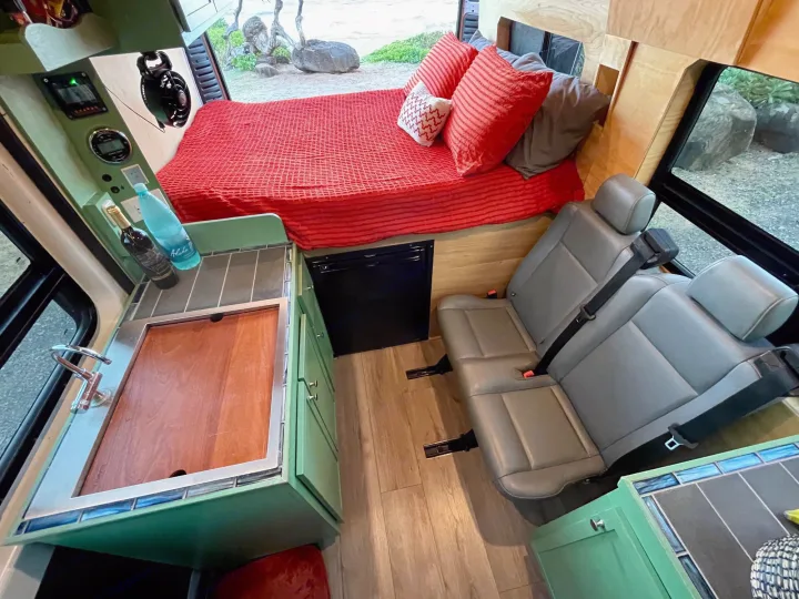 Inside this RV rental: a compact kitchen, comfortable bed with red bedding, and passenger seats. Ideal for road trips.. Dodge Ram Promaster 1500 2021