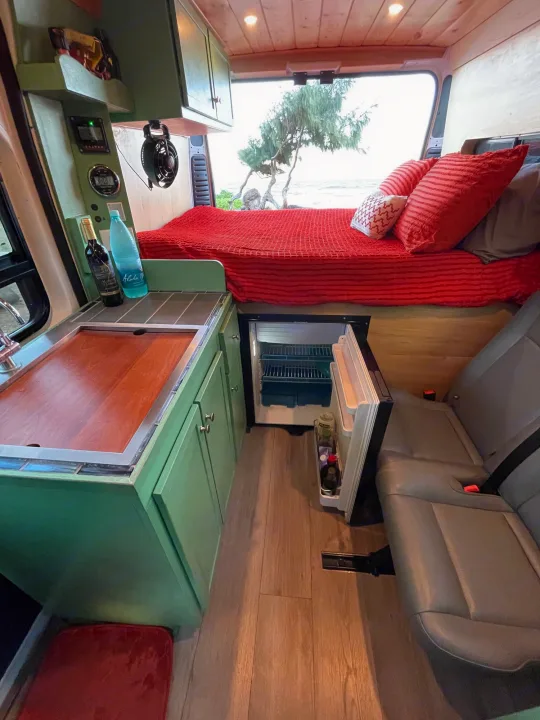 Inside a modern RV rental: green cabinets, kitchen, cozy red bed, fridge, and a back window view of the beach.. Dodge Ram Promaster 1500 2021