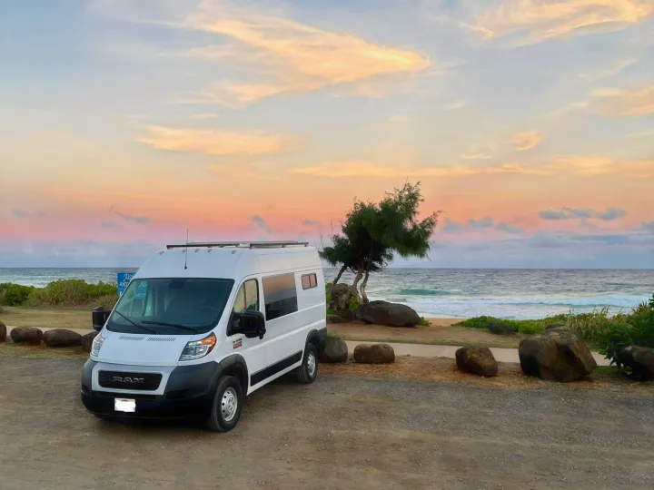 White rv rental camper van at a tropical beach sunset, with large rocks and ocean waves visible.. Dodge Ram Promaster 1500 2021