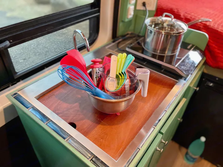 Compact RV rental kitchen features a sink, induction cooktop with a pot, and a bowl of colorful cooking utensils.. Dodge Ram Promaster 1500 2021