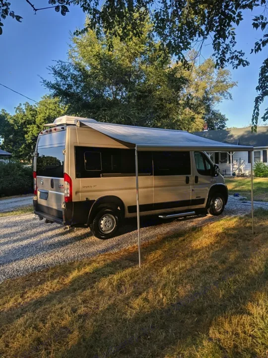 Light tan RV rental with extended awning on gravel driveway, surrounded by trees. Your next travel adventure awaits!. Roadtrek Roadtrek Class B 2017