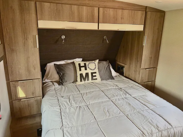 An RV rental bedroom setup, featuring a cozy bed with a "HOME" pillow, overhead cabinets, and side storage.. Grand Design Imagine 2021